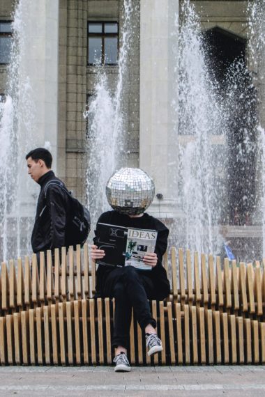 Mann mit Disko-Kugel auf dem Kopf - auf einer Bank sitzend - ein Magazin über innovative Ideen in der Hand