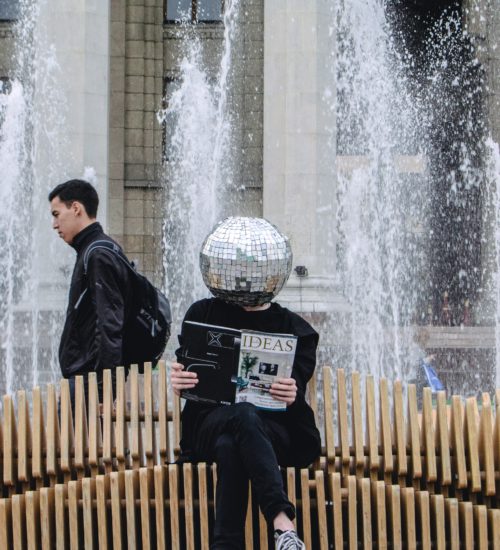 Mann mit Disko-Kugel auf dem Kopf - auf einer Bank sitzend - ein Magazin über innovative Ideen in der Hand